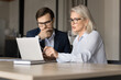 © fizkes - Serious younger and elder professional colleagues in glasses working together at laptop. Mature project manager, businesswoman showing online presentation on laptop to partner man