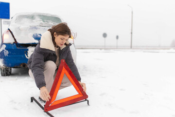 Wall Mural - woman puts up a warning triangle on a snowy road in winter, car breakdown in winter