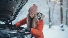 A young woman repairs a car in winter on the snow in the background. The foreground is a dead battery.