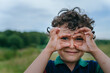Curly hispanic little boy in polo stands on meadow, against forest makes binoculars by hands looks at camera, smiles, enjoying leisure outdoors. Childhood, healthy children.