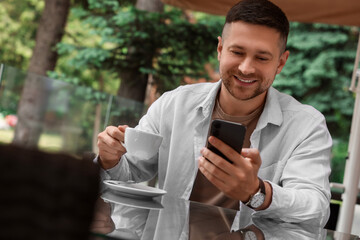 Wall Mural - Handsome man with cup of drink sending message via smartphone at table in outdoor cafe