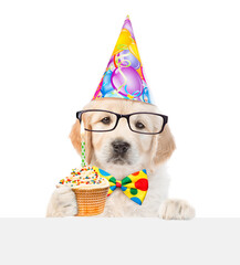 Poster - Golden retriever puppy wearing eyeglasses, tie bow and party cap looks above empty white banner and shows cupcake with burning candle. isolated on white background
