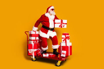 Wall Mural - Full body profile portrait of charismatic aged santa ride shop trolley pile stack christmas giftbox isolated on yellow color background