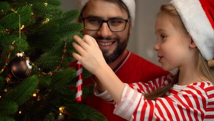 Wall Mural - Close up of father and daughter in red santa hats fun decorate Christmas tree with cane lollipop at home. Happy family celebrate New New Year and Christmas