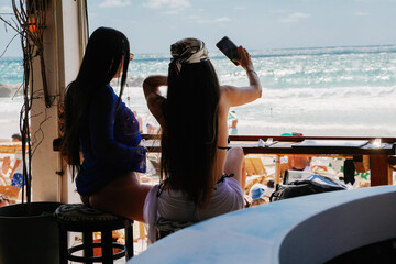 2 sexy womens sitting on the bar at beach restaurant while summer time and take selfie infant of the ocean view