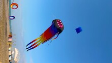 Vertical Video: Colorful Kites Are Flying In The Clear Blue Sky And Swaying In The Wind During Summer Outdoor Festival Near The Sea. Childhood, Freedom And Leisure Time Concept