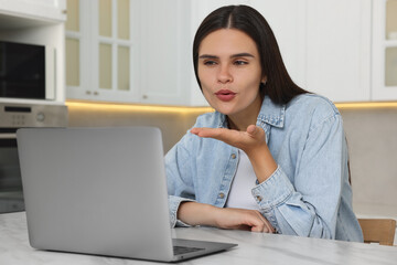 Poster - Happy young woman having video chat via laptop and blowing kiss at table in kitchen