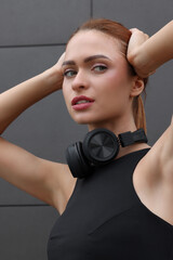 Wall Mural - Beautiful woman in stylish gym clothes with headphones posing near dark grey wall on street, closeup