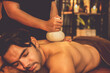 Hot herbal ball spa massage body treatment, masseur gently compresses herb bag on man body. Tranquil and serenity of aromatherapy recreation in warm lighting of candles at spa salon. Quiescent