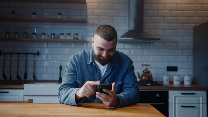 Young adult man sitting against the kitchen counter checking messages read notice receive good news on smartphone in e-mail celebrate great opportunity