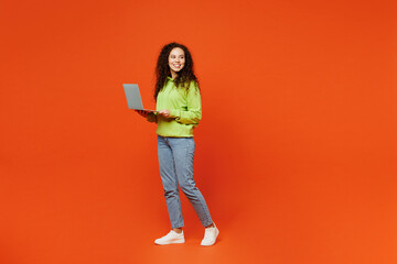 Wall Mural - Full body young IT woman of African American ethnicity she wears green hoody casual clothes hold use work on laptop pc computer look aside isolated on plain red orange background. Lifestyle concept.