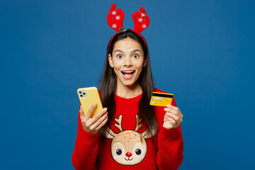 Wall Mural - Merry amazed young Latin woman wear red Christmas sweater decorative deer horns on head posing hold credit bank card mobile cell phone isolated on plain blue background Happy New Year holiday concept.