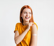 © kucherav - Portrait of a red-haired smiling young woman after getting a vaccine standing on a white background, showing her arm with bandage after receiving vaccination