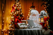 Santa Claus reads letters from children in his home at the table against the backdrop of Christmas lights.