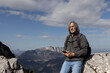 © Guzel - Handsome gray-haired man with long hair sitting with phone in hands on high slope against bright sky, Austria