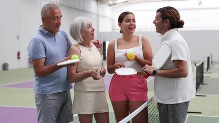 Wall Mural - Happy smiling men and women of different ages in sportswear with rackets and balls in hands talking friendly near net on indoor pickleball court. High quality 4k footage