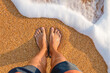 male feet barefoot on the waves of sea foam on a sandy golden beach on a summer day. top view of male legs in shorts and clear waters of the ocean