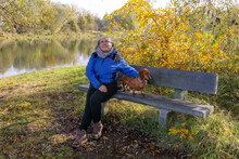 Senior Hiker Taking A Break, Sitting On Wooden Bench With Her Dog, River Surrounded By Autumn Trees In Background, Belgian Nature Reserve De Wissen Maasvallei, Sunny Day In Dilsen-Stokkem, Belgium
