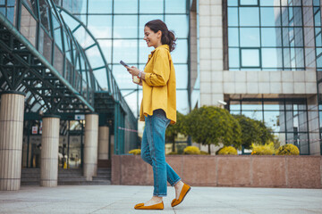 Wall Mural - Woman texting and drinking coffee outdoors. Young smiling brunette woman holding a smartphone and a cup of coffee looking at device screen against the background of an office building.