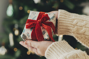 Wall Mural - Merry Christmas! Woman in cozy sweater holding stylish christmas gift in festive wrapping paper with bow on background of christmas tree lights. Atmospheric winter holidays. Happy holidays!