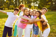 Children Have Fun And Make Friends At The Summer Camp. Group Of Kids Spending Time Outdoors Together. Bunch Of Happy Girls And Boys Hugging Each Other In A Green Sunny Park