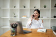 © Wasana - Happy Asian woman talking on the phone while sitting in the office.