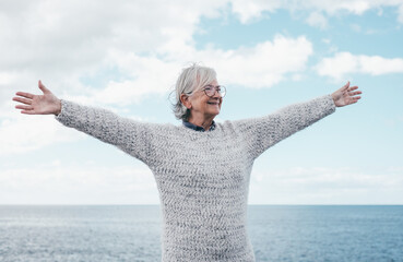 Wall Mural - Smiling caucasian elderly woman with open arms in travel, retirement o vacation at sea enjoying nature and freedom. Horizon over water