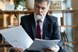 © opolja - Mature businessman in formal suit concentrating on reading the report, sitting in the lobby and preparing for the meeting.