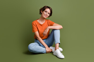 Full size photo of smart good mood woman dressed orange t-shirt jeans in glasses sit on floor smiling isolated on khaki color background