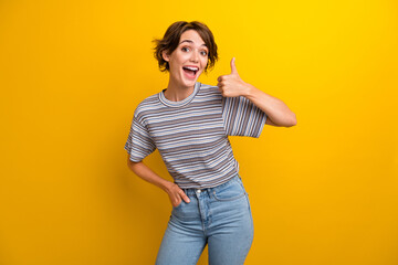 Poster - Portrait of overjoyed lovely funky girl wear grey t-shirt showing thumb up holding hand in pocket isolated on yellow color background