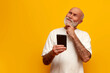 pensive old bald grandfather in white t-shirt uses smartphone and thinks on yellow isolated background