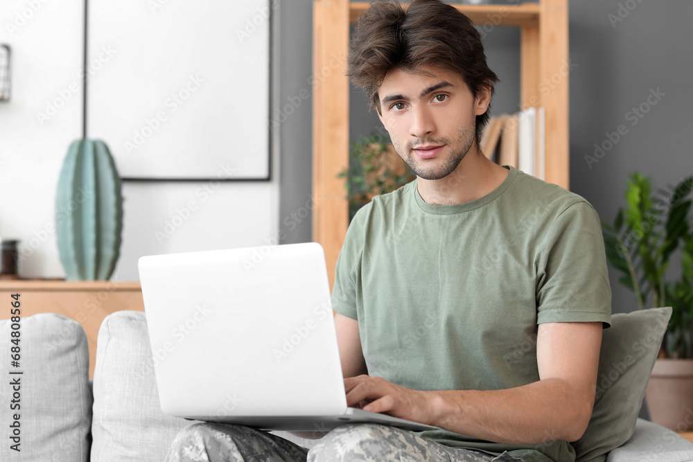 Young male soldier with laptop sitting on sofa at home