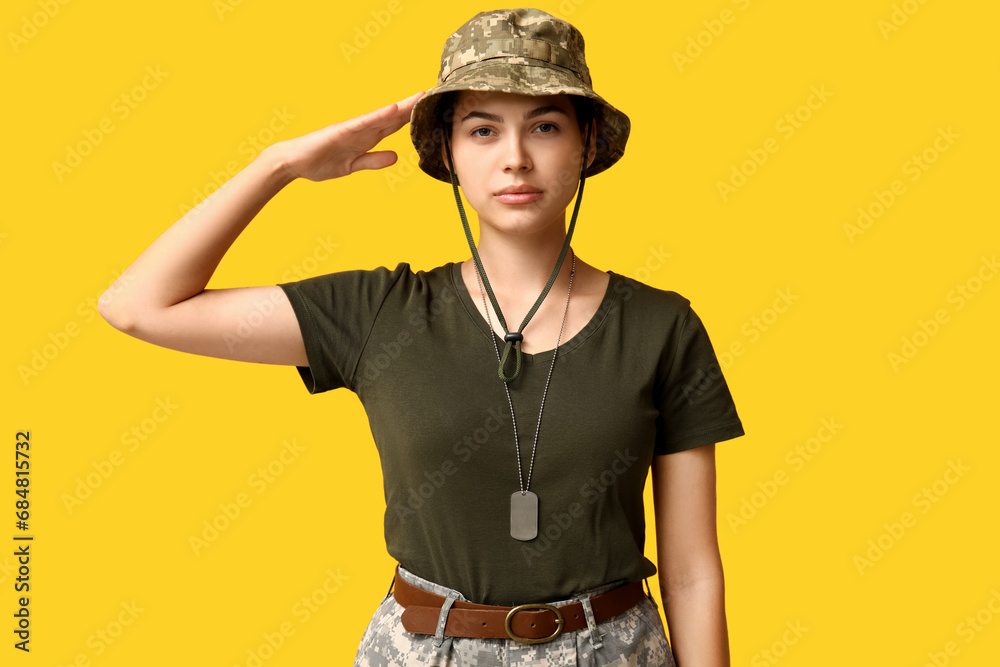 Confident young female soldier in uniform saluting on yellow background
