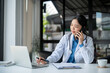 © bongkarn - A doctor is talking on the phone with her patient while working in her hospital office.