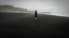Young Beautiful Woman In Black Dress Walking On Black Sand Beach Iceland, Misty Dramatic Mountain Landscape, Tracking Shot
