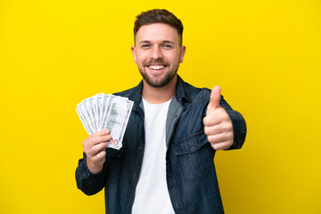 Wall Mural - Young caucasian man taking a lot of money  isolated on yellow background with thumbs up because something good has happened