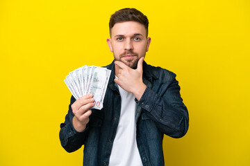 Wall Mural - Young caucasian man taking a lot of money  isolated on yellow background thinking