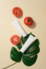 Wall Mural - On a beige background, set of cosmetic tubes displayed with ripe tomatoes and green monstera leaf. Advertising scene. Tomatoes contain compounds that help shrink pores and brighten skin tone.