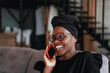 © Iona - excited young African girl in glasses and traditional clothes in black turban is talking on the phone sitting on the sofa at home against  background of a blurry interior with a staircase. Real people