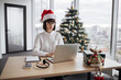 © sofiko14 - Caucasian businesswoman wearing glasses sitting in office near xmas tree and working on laptop during the holiday season, typing on computer keyboard.