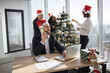 © sofiko14 - Caucasian businessman in suit and Santa hat talking on smartphone while using portable laptop computer looking at window on background of colleagues decorating Christmas tree.