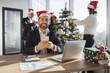 © sofiko14 - Businessman in Santa hat sit at table and using smartphone in decorated festive office looking at camera. Male freelancer taking break during working process while coworkers decorating Christmas tree