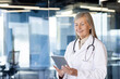 © Liubomir - Close-up photo. Smiling senior female doctor, nurse standing in clinic office and using tablet, searching, typing, consulting online.