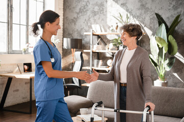 Closeup image of caregiver nurse caretaker doctor greeting giving handshake to senior old elderly grandmother patient female with walking frame with motion immobility in hospice at home. Homecare