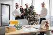 © sofiko14 - Multicultural team of business people celebrating new year in office with panoramic windows, decorating Christmas tree together, preparing for holiday party.