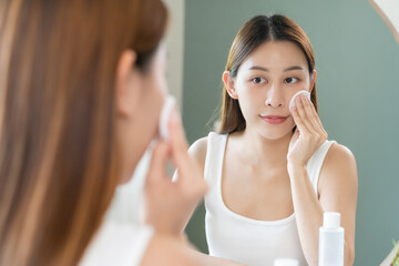 Wall Mural - Happy beauty, attractive asian young woman, girl looking reflect in mirror, hand holding cotton pad, applying facial wipe on her face remover makeup, essence or lotion for treatment, skin care routine