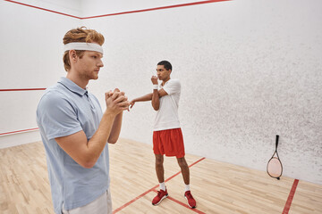 Wall Mural - young redhead man with headband warming up near african american friend inside squash court