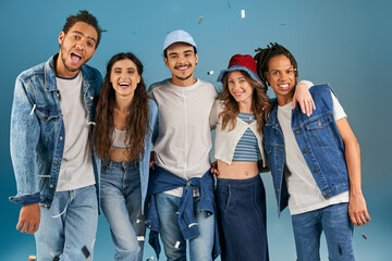 cheerful interracial friends in trendy casual attire looking at camera under confetti rain on blue