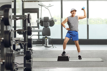 Wall Mural - Elderly man in sportswear exercising with a step aerobic platform and a small weight at a gym