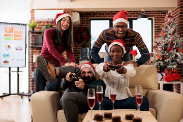 Wall Mural - Excited colleagues team in santa hats participating in video gaming competition in decorated workplace. Cheerful coworkers celebrating christmas and playing console game together